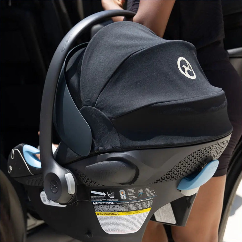 Close-up of the Cybex Aton G Swivel Sensorsafe Infant Car Seat in black fabric with blue accents, parent carrying it toward a vehicle.