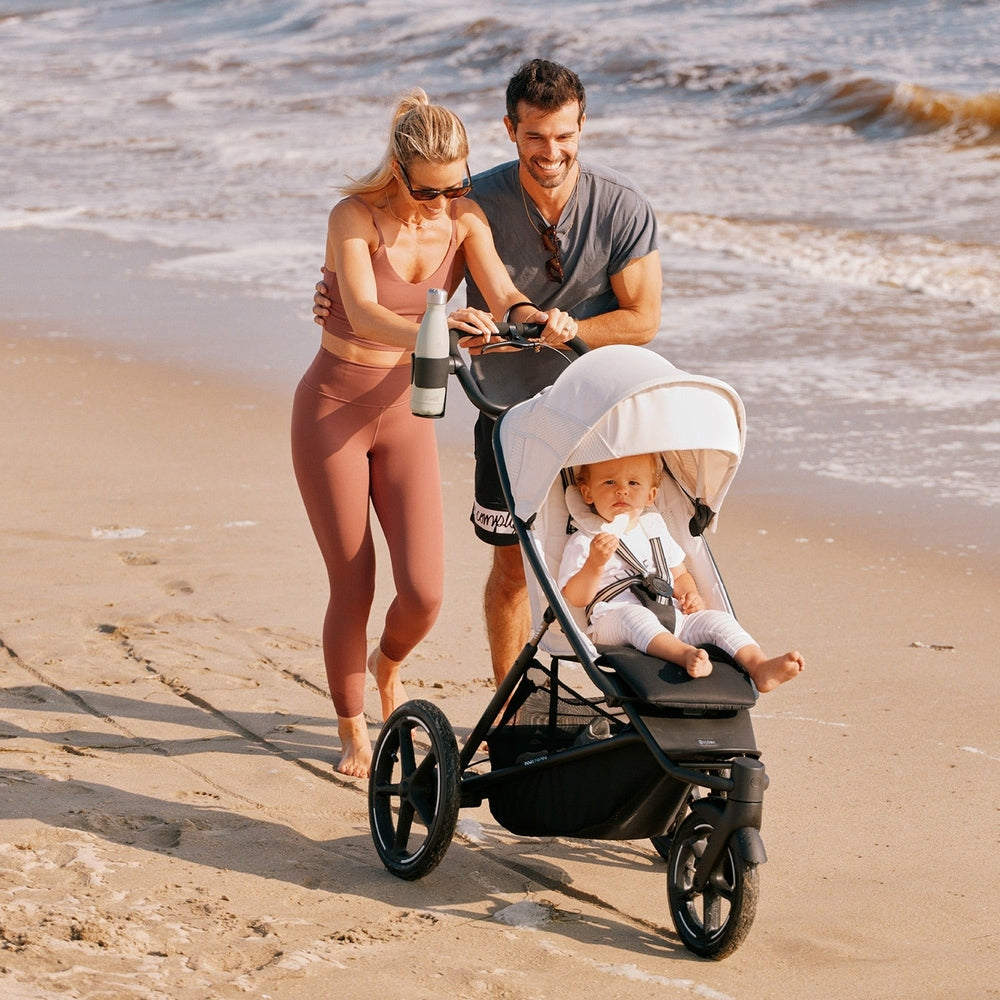 Lifestyle image of the Cybex Avi Spin All Terrain Stroller on sunny beach; couple push a baby in the white-canopy stroller.