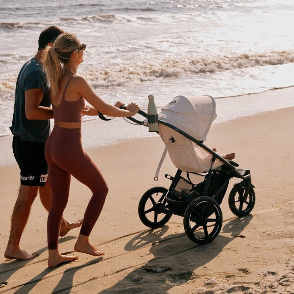 Close-up of the Cybex Avi Spin All Terrain Stroller pushed along sandy beach by a couple at sunset, white canopy, rugged wheels.