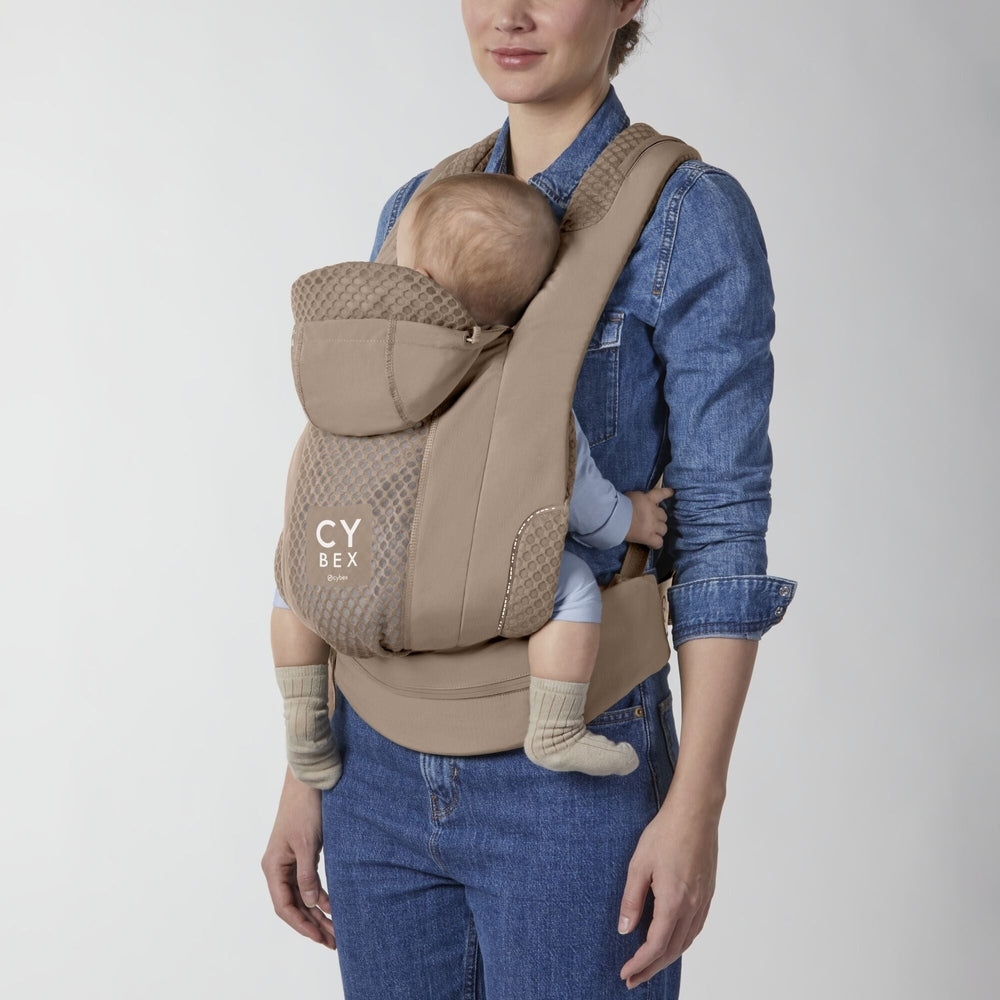 Close-up of the Cybex Coya Carrier beige cybex coya baby carrier with honeycomb mesh panels and hood, worn by a person in denim; baby nestled inside.