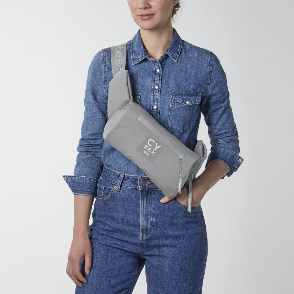 Lifestyle image of the Cybex Coya Carrier grey cybex coya baby carrier with logo patch, worn crossbody over a denim shirt.