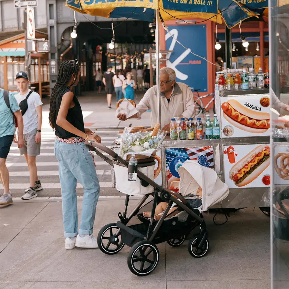 Lifestyle image of the Cybex Gazelle S V2 Stroller in black with bassinet, parent pushes baby beside a busy street hot dog cart.