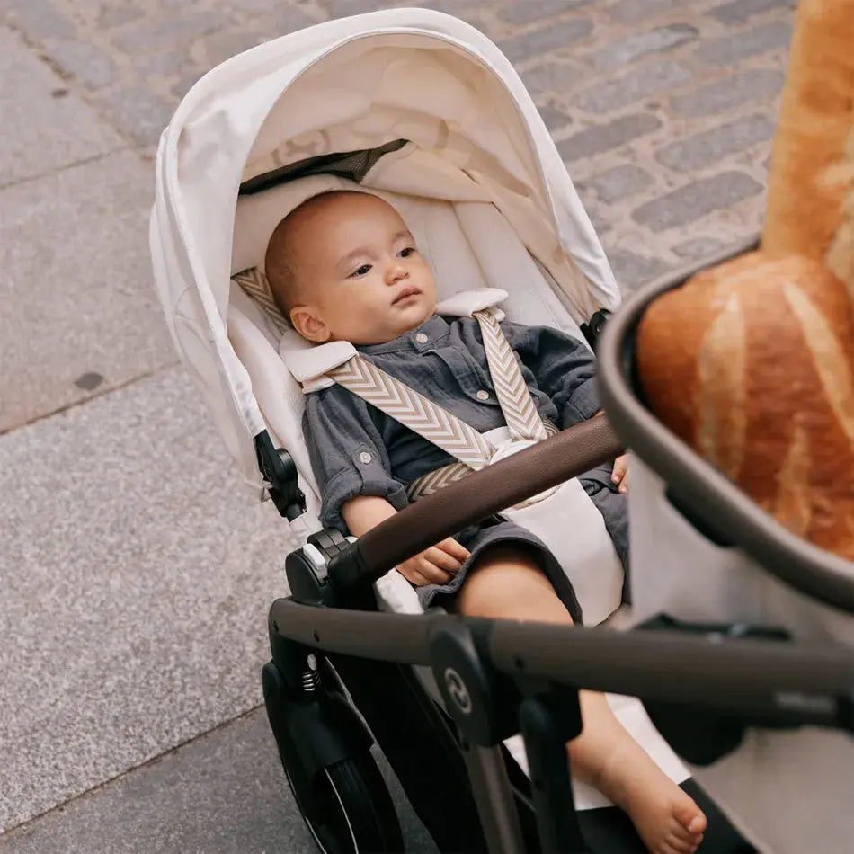 Lifestyle image of the Cybex Gazelle S V2 Stroller ; white canopy, grey seat, beige striped harness, with a baby securely seated on a sidewalk.