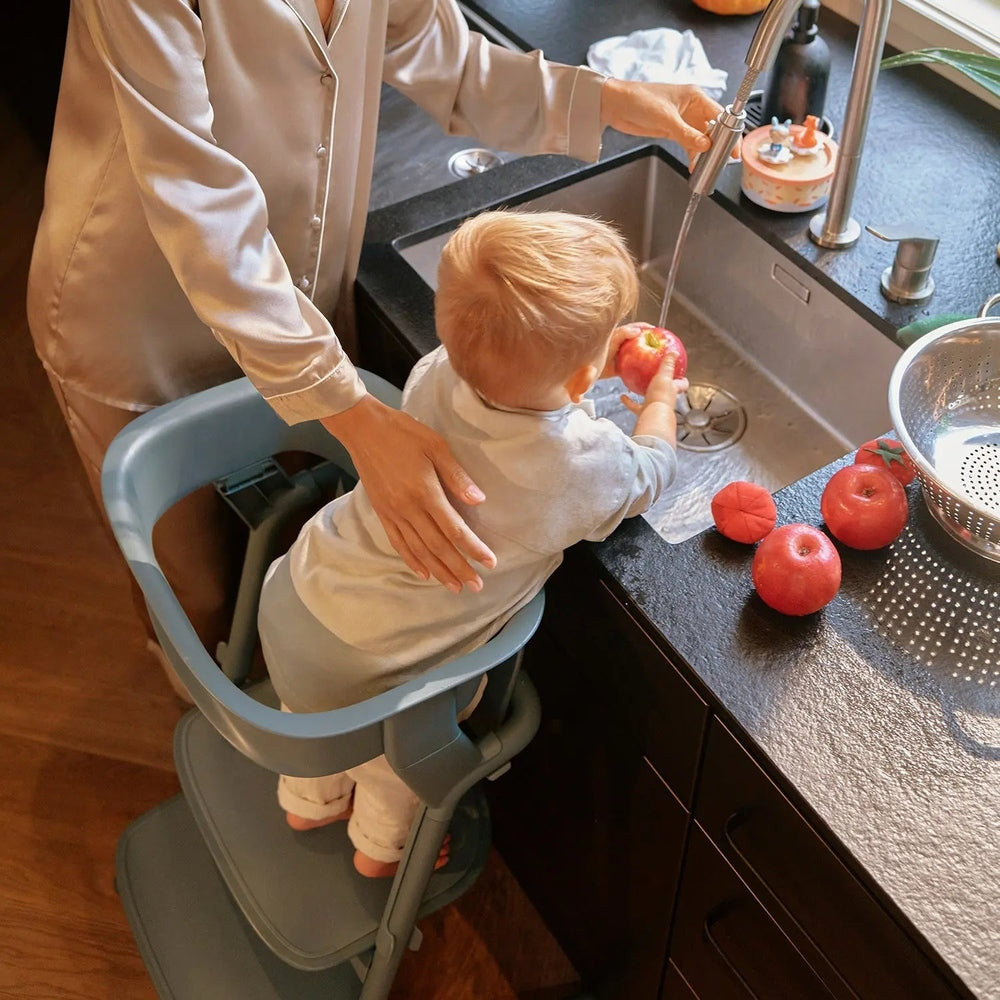Lifestyle image of the Cybex Lemo Training Tower high chair in blue-gray, toddler at the kitchen sink with a parent helping wash an apple.