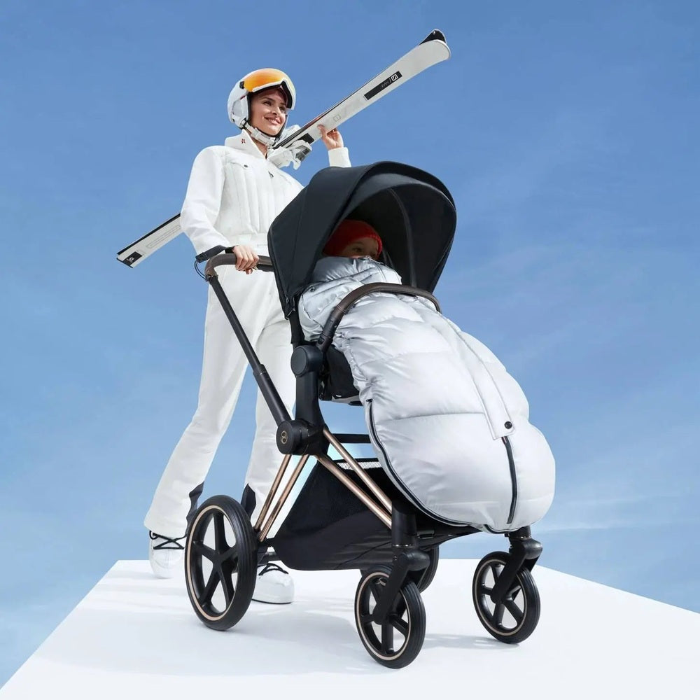 Lifestyle image of the Cybex Winter Footmuff on a stroller, silver-gray quilted cover keeping a child warm during a bright winter stroll.