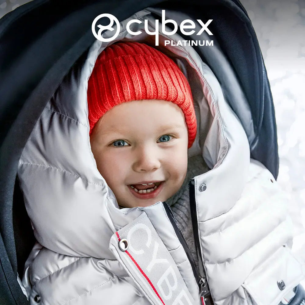 Lifestyle image of the Cybex Winter Footmuff in platinum, shown snugly around a smiling baby in a stroller with a red knit hat.