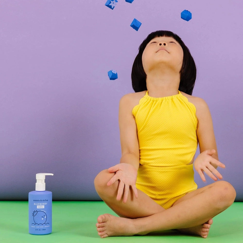 Dabble & Dollop All-Natural Shampoo, Body Wash & Bubbles Blueberry bottle beside a child in a yellow outfit with blue bubbles overhead.