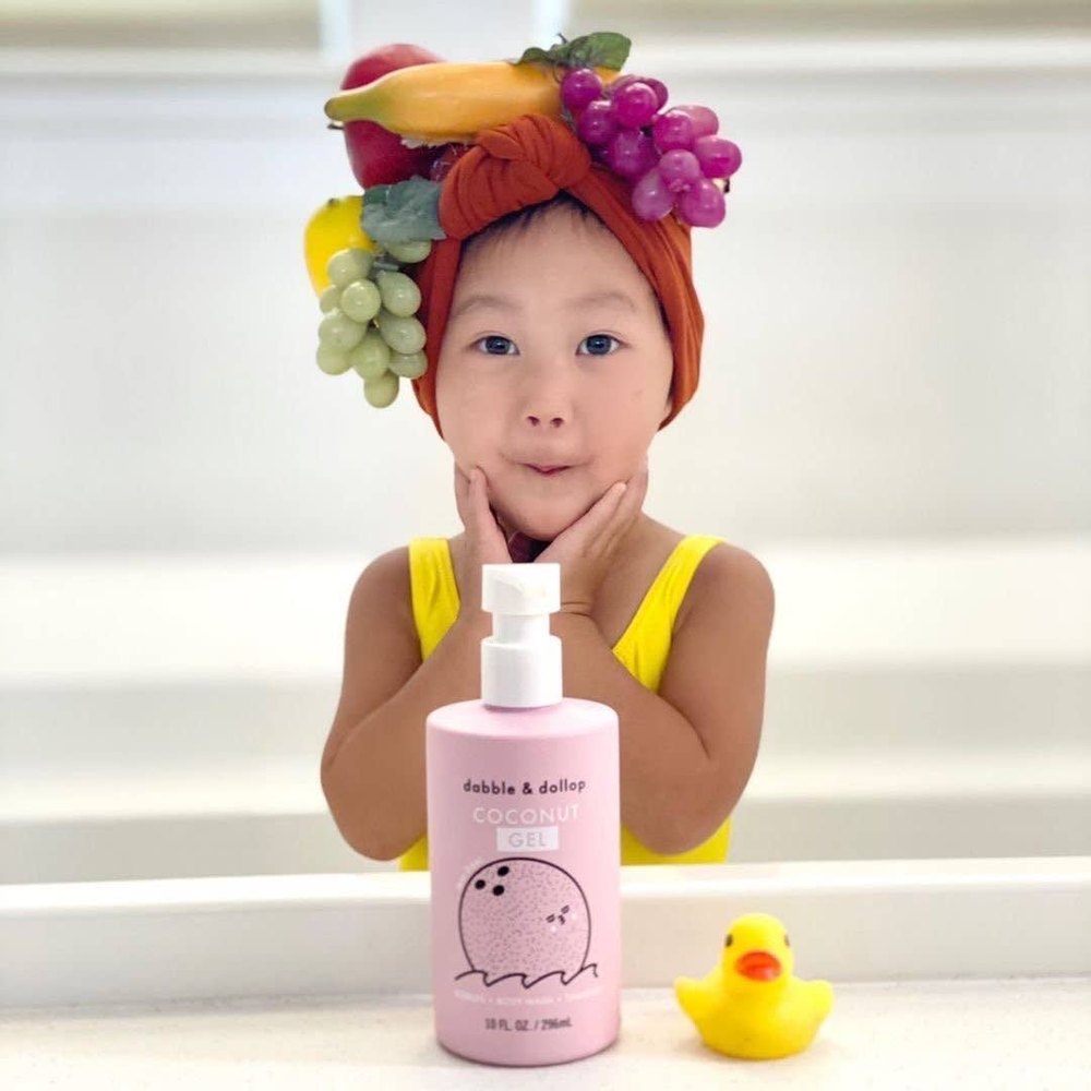 Dabble & Dollop All-Natural Shampoo, Body Wash & Bubbles Coconut bottle with pump on counter beside a yellow rubber duck, child smiles nearby.