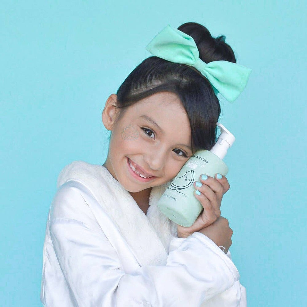 Dabble & Dollop All-Natural Shampoo, Body Wash & Bubbles Honeydew bottle held by a smiling girl in a white robe.