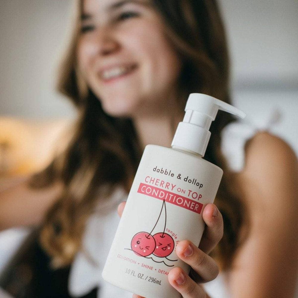Dabble & Dollop Cherry on Top Hair Conditioner bottle with pump held by a smiling woman, pink cherries illustration.