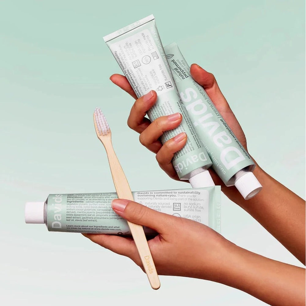 DAVIDS Toothpaste Fluoride Free Peppermint tubes, mint-green packaging, two hands squeezing a tube beside a wooden toothbrush.