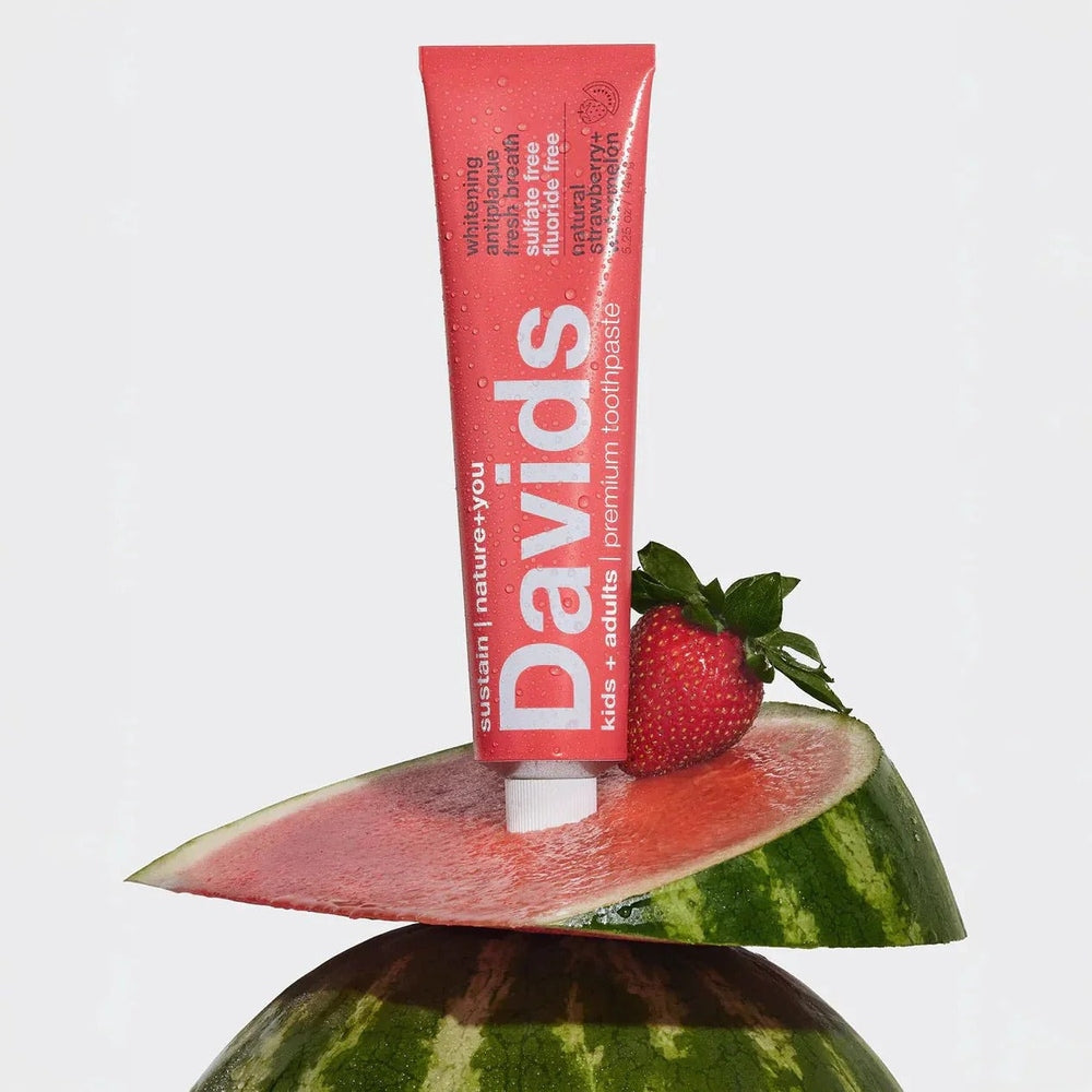 DAVIDS fluoride-free strawberry watermelon toothpaste tube upright on a watermelon slice beside a strawberry.