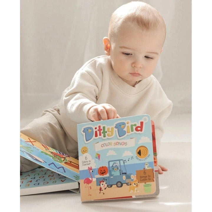 Ditty Bird Sound Book Color Songs USB-C Rechargeable, a baby taps the colorful pages of the blue cover.