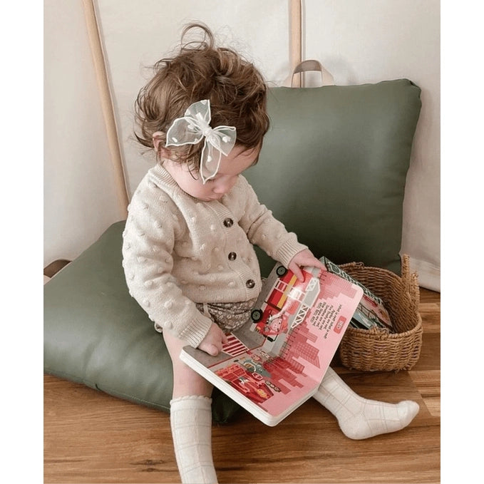 Ditty Bird Color Songs USB-C Rechargeable toy; toddler sits reading a colorful book with a white bow and cream cardigan.