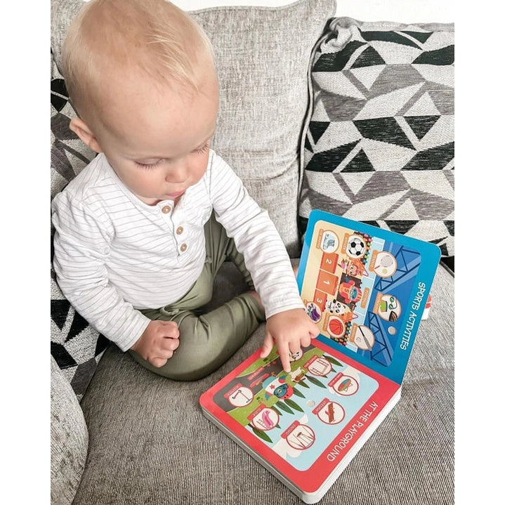 Ditty Bird Sound Book First 100 Places USB-C Rechargeable; baby taps a colorful page on the open book beside a gray sofa.