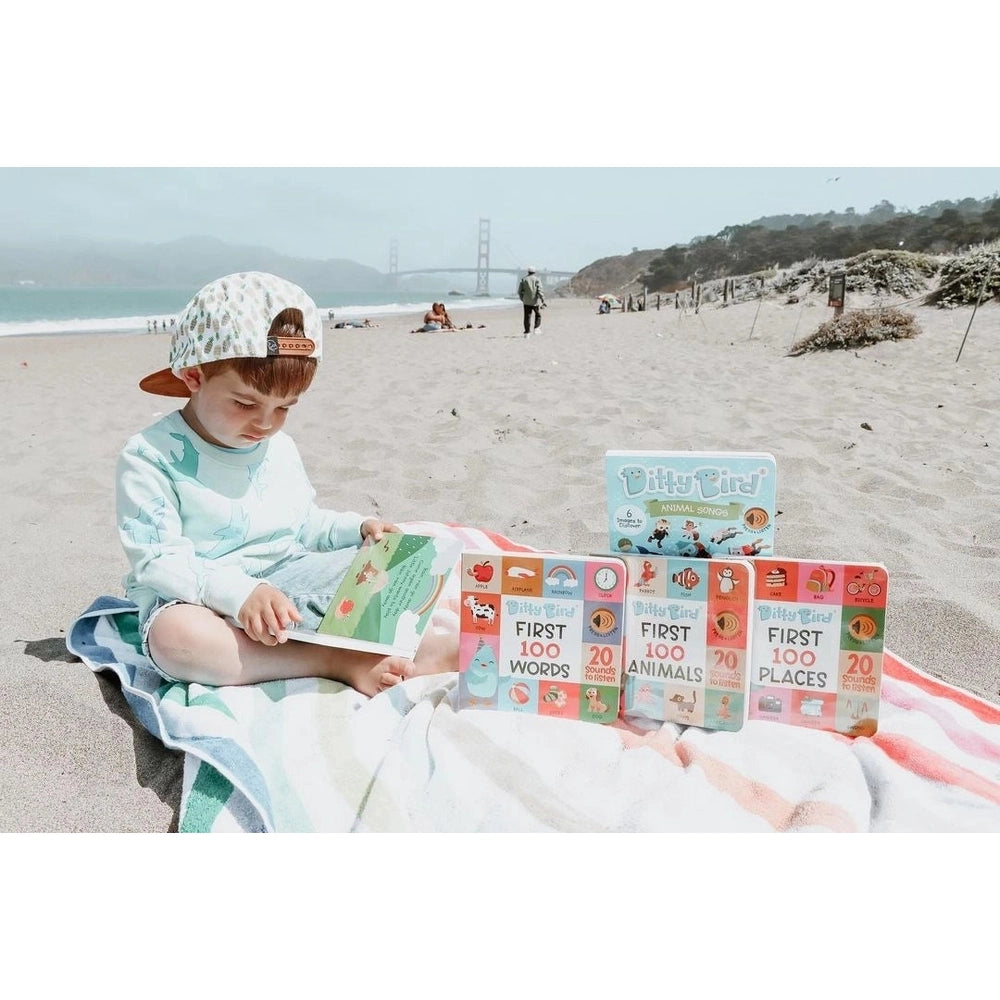 Ditty Bird Sound Book First 100 Places USB-C Rechargeable being read by a child on a sunny beach beside colorful volumes.