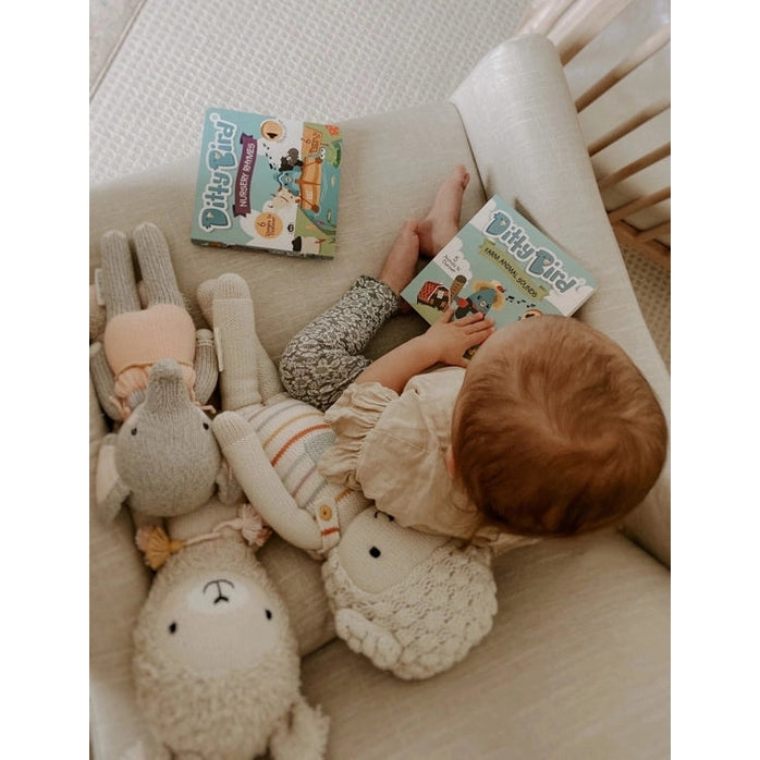 Ditty Bird Sound Book Nursery Rhymes USB-C Rechargeable held by baby in a cozy crib with plush toys nearby.