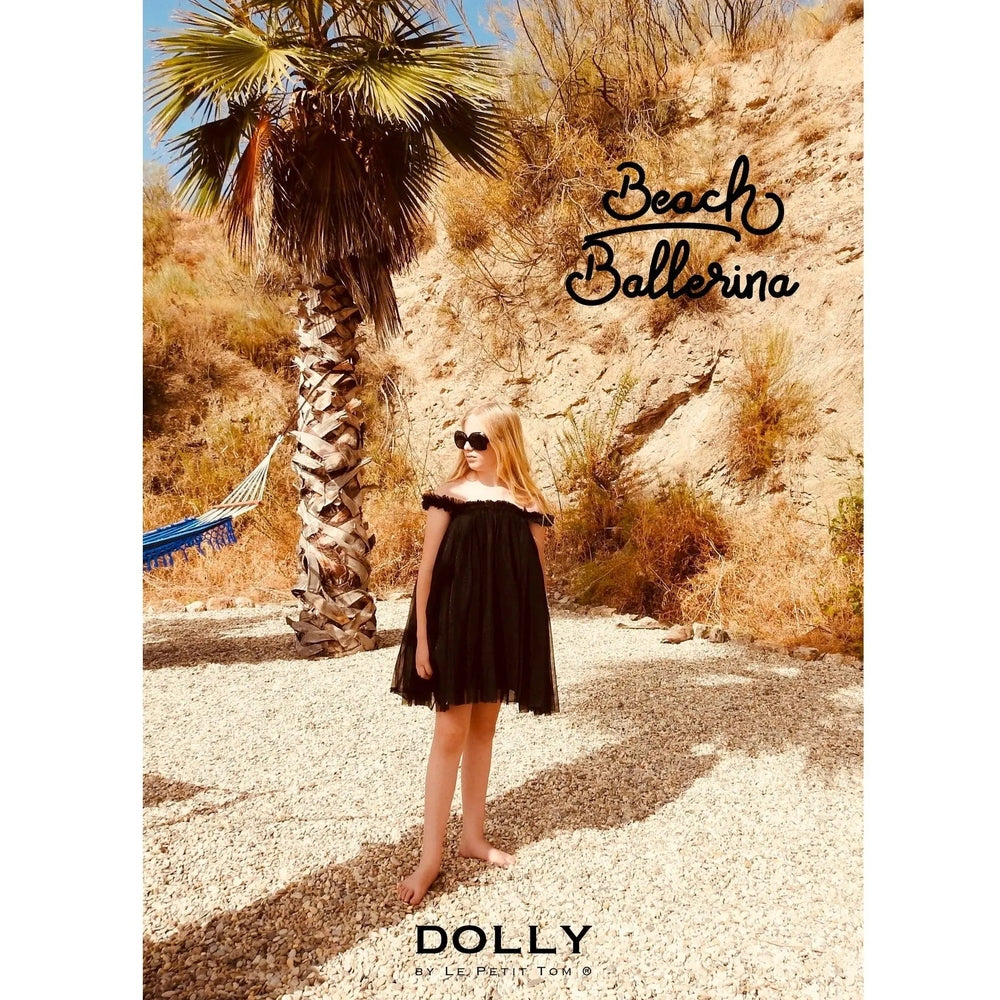 Dolly By Le Petit Tom Tutu Dress Beach Cover Up Little Black worn by a barefoot toddler on a sunny pebble beach.