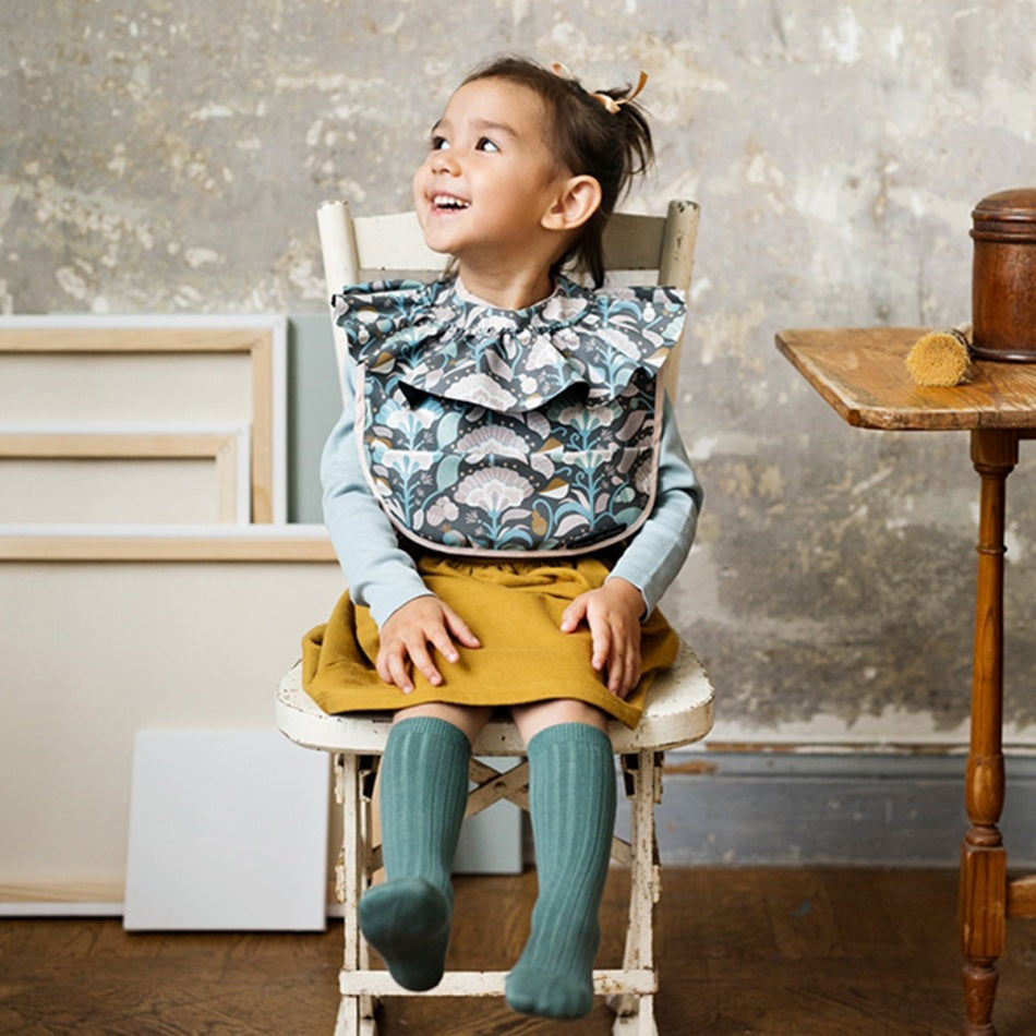 Elodie Details Baby Bib Midnight Bells in blue-gray floral print worn by a cheerful toddler.