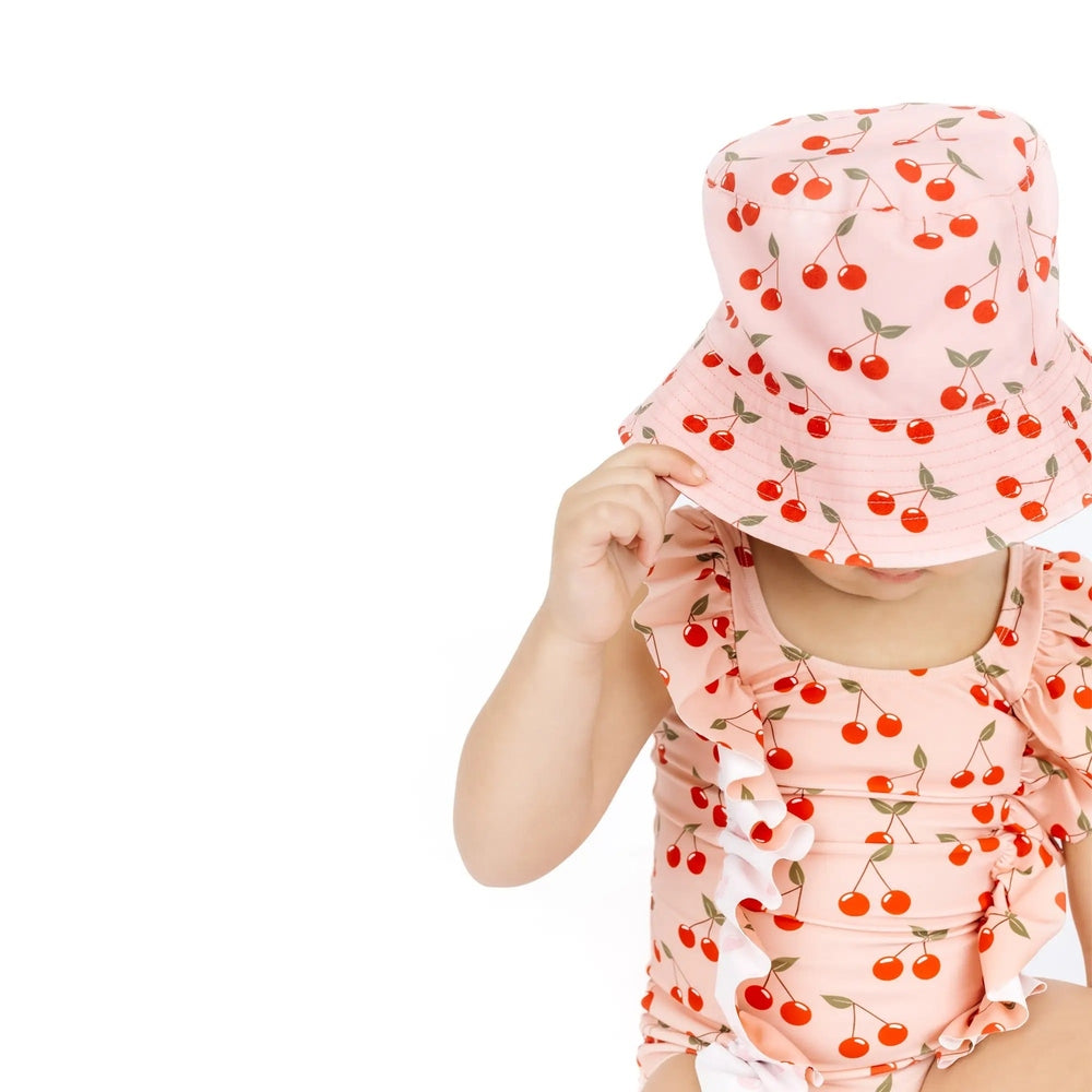 Child wearing a cherry-patterned outfit with a matching hat on a white background