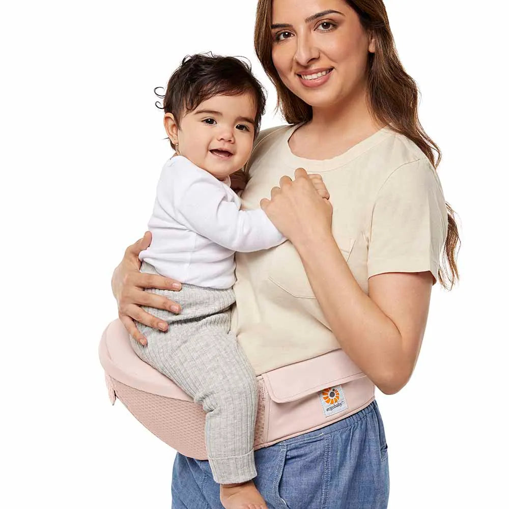 Ergobaby Alta Hip Seat Baby Carrier with pink waistband worn by mother carrying a smiling baby in a white onesie.