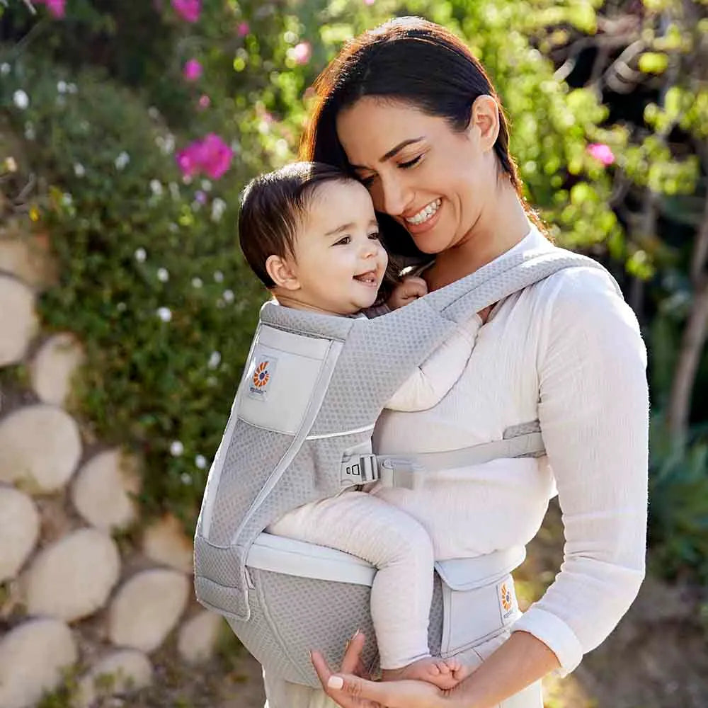 Ergobaby Alta Hip Seat Baby Carrier in light gray worn by a smiling mom outdoors, with baby securely on hip.