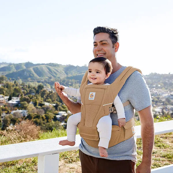 ERGOBABY Alta Hip Seat Baby Carrier in tan worn by dad outdoors with a smiling baby secured.