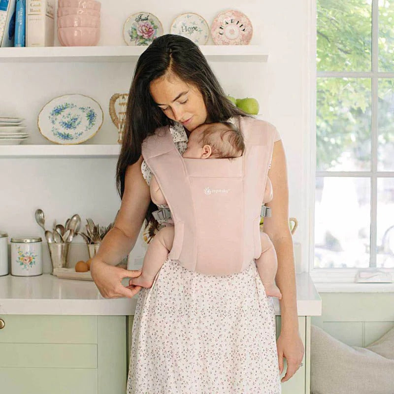ERGOBABY Embrace Newborn Carrier Blush Pink worn by mom carrying a sleeping infant in a cozy kitchen.