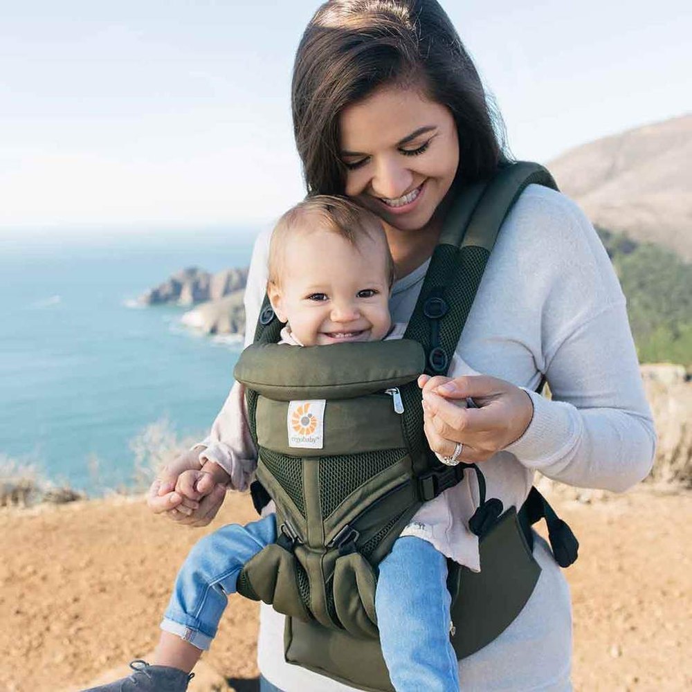 Ergobaby Omni 360 Cool Air Mesh olive carrier worn by a smiling mom outdoors by the coast, with a securely seated baby.