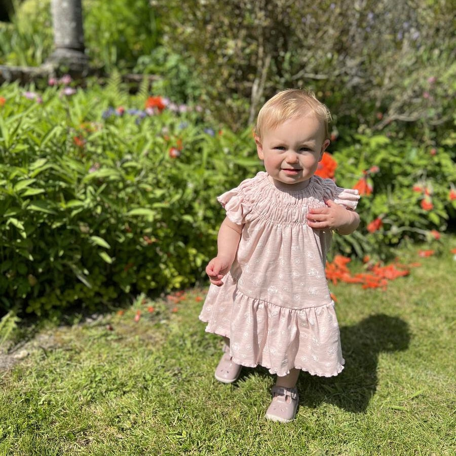 Ettie & H Pale Pink Flowers Dress; a baby girl stands in a sunny garden wearing a pink floral dress with ruffled sleeves.