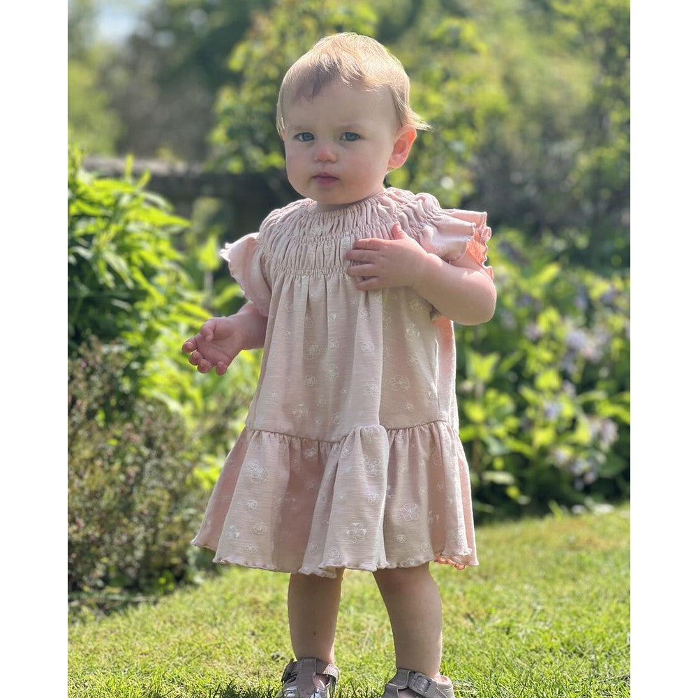 Ettie & H Pale Pink Flowers Dress; baby stands in a sunlit garden, showcasing a smocked bodice and ruffled sleeves.