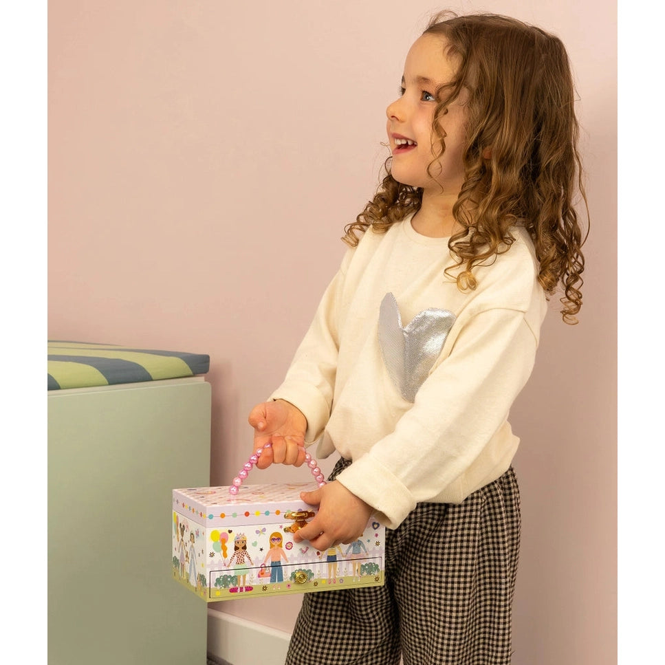 Joyful child in a cream top holds Floss And Rock Musical Jewellery Box Best Friends with a pink beaded handle.