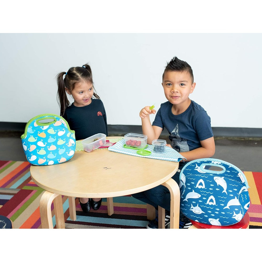 Blue shark-pattern insulated lunch bag with a handle, and turquoise whale-pattern bag on a round table as two kids eat.