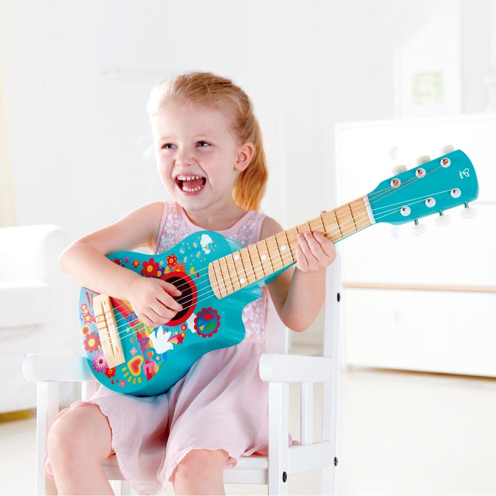 Hape Flower Power Guitar, turquoise with colorful flowers, child sits on white chair strumming the toy guitar.