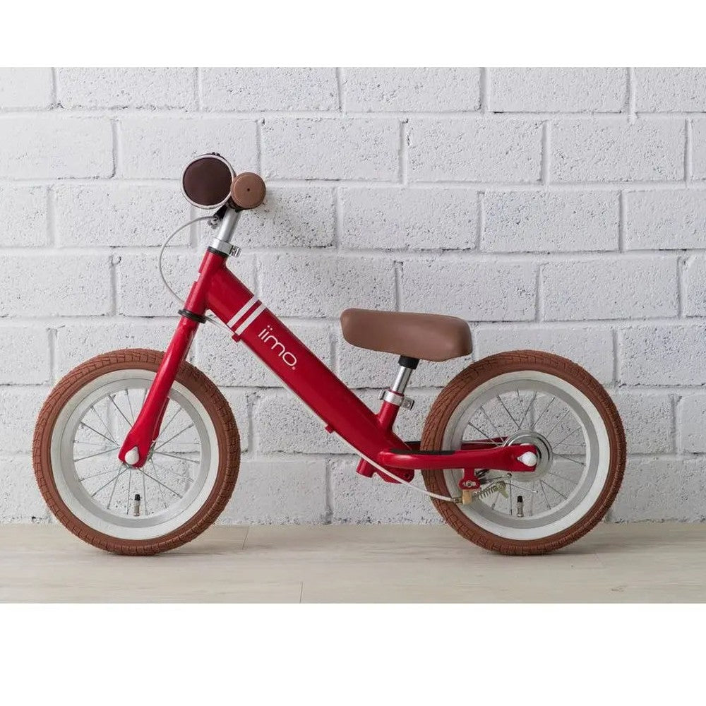 Iimo Balance Bike red frame with brown saddle and tires, shown ready for toddlers against a white brick wall.