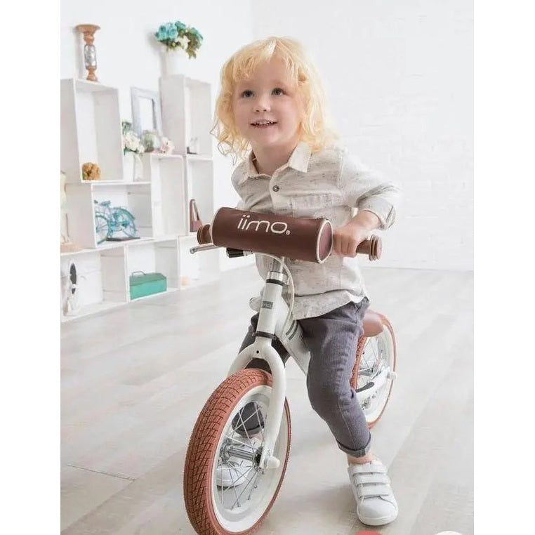Iimo Balance Bike for toddlers; white frame, brown padded handlebars, red tires, child rides indoors smiling.