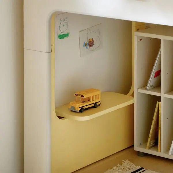 Iloom Cabin Hanging Shelf in pale yellow, mounted beside a crib with a toy bus on its curved shelf.