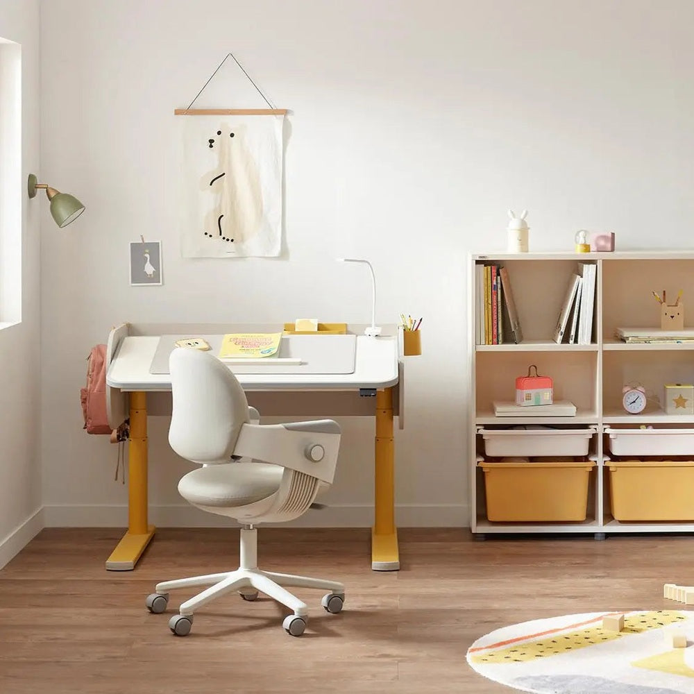 Iloom Jerome Motion Desk white with yellow legs; height-adjustable frame shown beside a white ergonomic chair in a bright room.