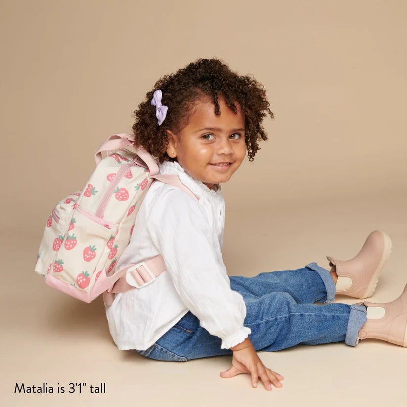 Itzy Ritzy Backpack, pink strawberry print with light pink straps, worn by a smiling girl as she sits.