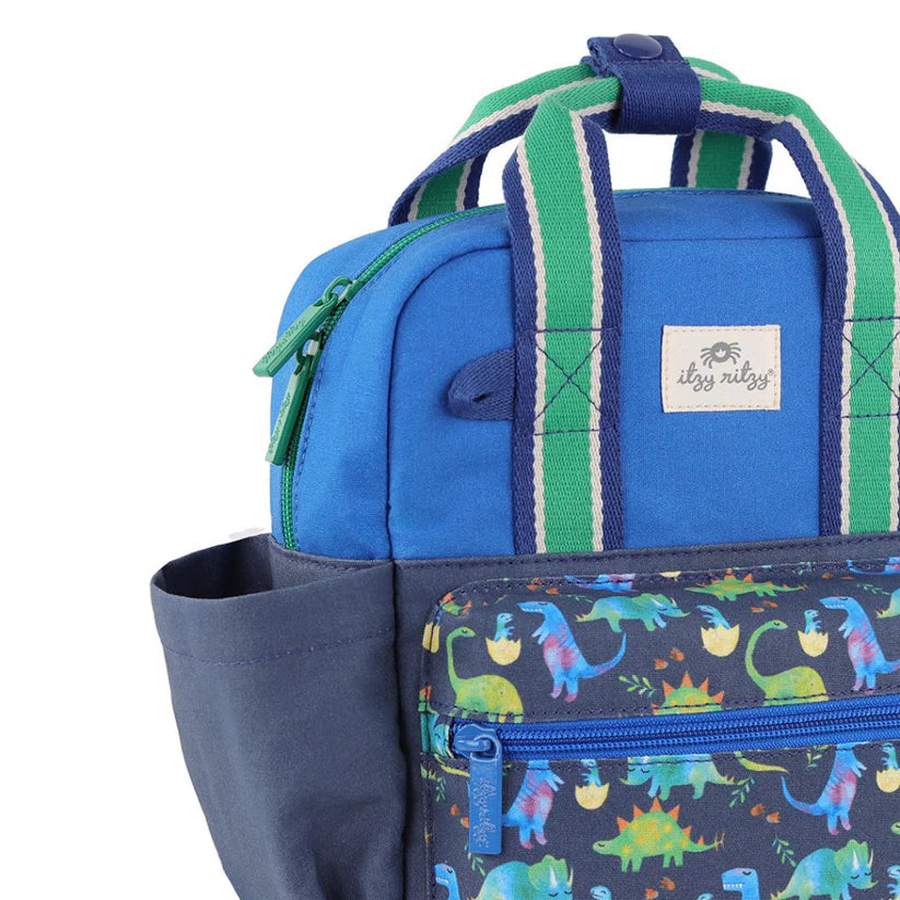 Itzy Ritzy Backpack in blue with a dinosaur-print front pocket and green striped handles ready to carry.