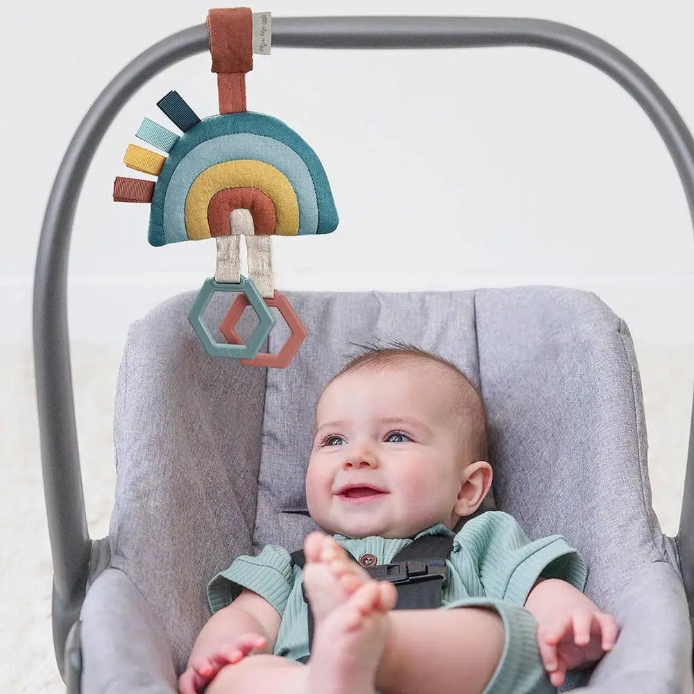 Itzy Ritzy Bitzy Bespoke Ritzy Jingle Attachable Travel Toy, rainbow felt with shapes, clipped to a car-seat handle as baby smiles.