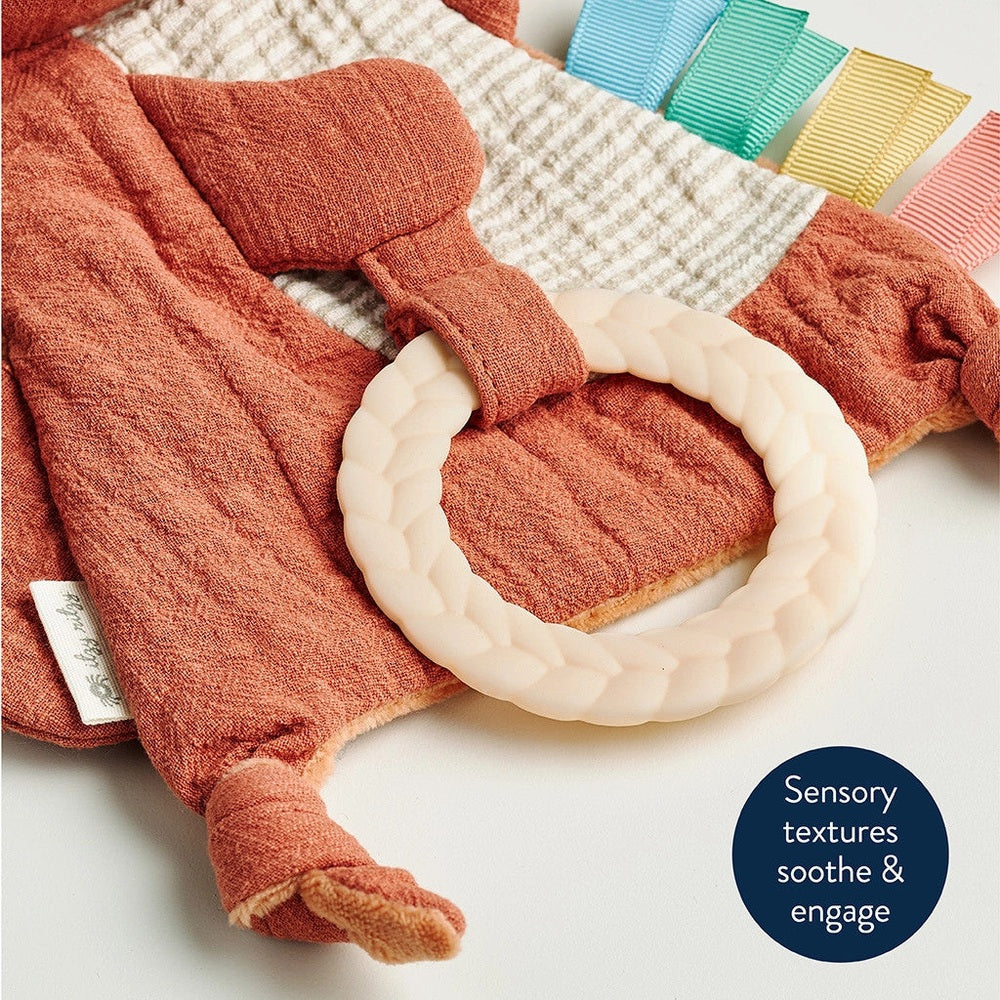 Itzy Ritzy Bitzy Crinkle sensory toy with teether, ivory braided ring on coral plush blanket with pastel ribbon tabs
