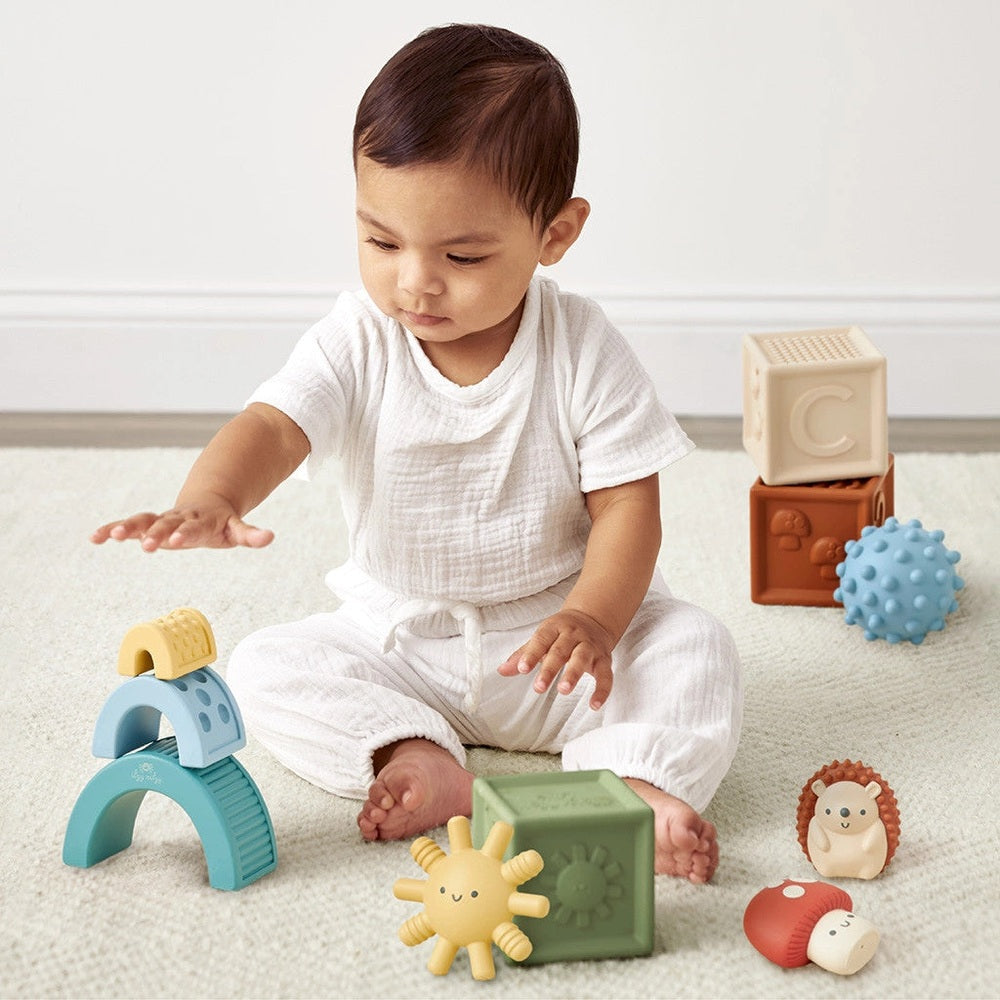 Seated baby reaching toward blue curved pieces with blocks and toy figures spread on a cream rug