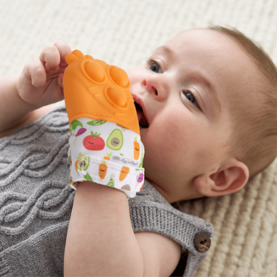 Itzy Ritzy Mitt Pop teething mitten with orange silicone grip; baby gnaws the fruit-printed fabric while lying on a rug.