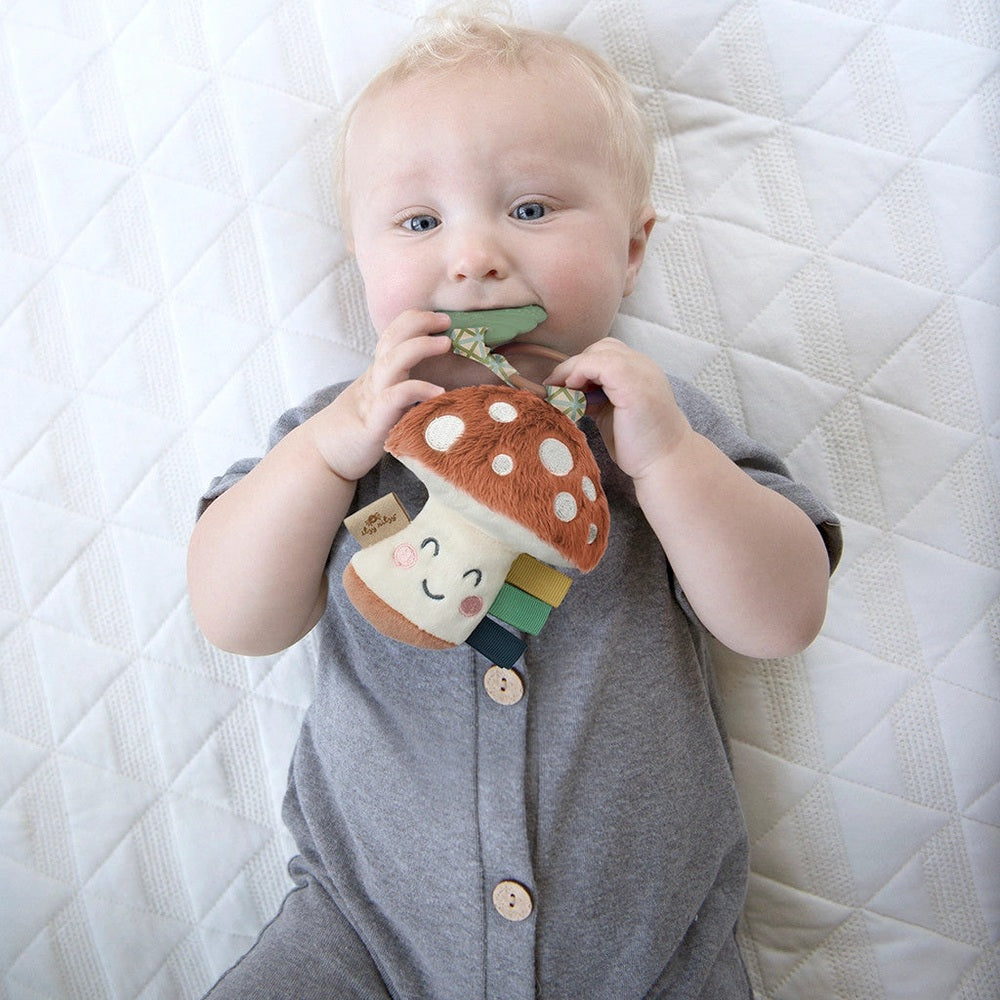 Itzy Ritzy Pal plush teether mushroom toy with colorful ribbon tabs, held by a baby in a gray onesie.