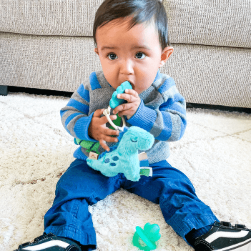 Itzy Ritzy Pal plush and teether toy, teal; baby sits on white carpet with teether and plush toy
