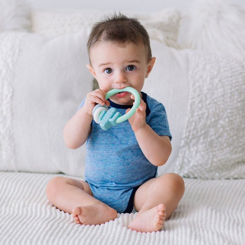 Itzy Ritzy Rattle Silicone Teether, mint green ring with light blue teething pieces; baby sitting on a bed.