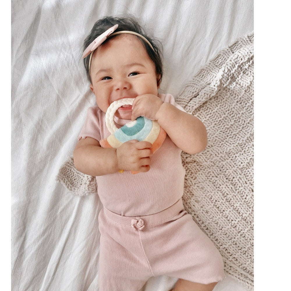Itzy Ritzy Ritzy Rattle Pal plush rattle teether in pastel rainbow colors, held by smiling baby in pink.