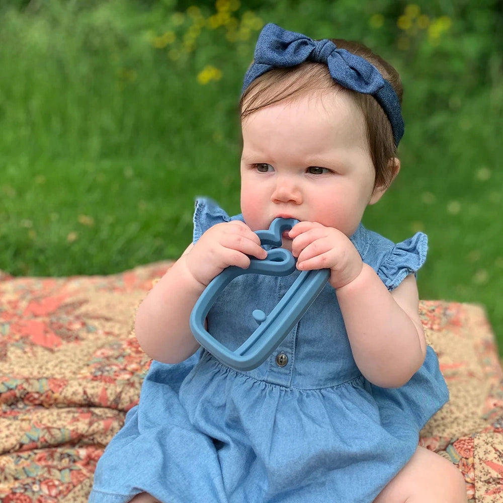 Itzy Ritzy silicone teether in blue, held by a baby in a denim dress and matching headband
