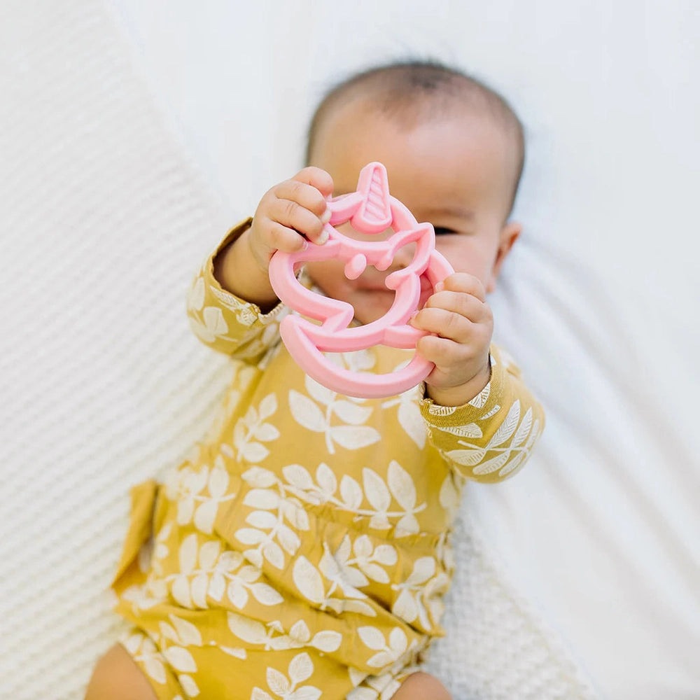 Itzy Ritzy silicone unicorn teether in pink, held by baby in yellow leaf-print outfit